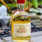 Flaske med julemjød, 35 cl, med rød forsegling placeret på en sten udendørs ved en vandfontæne.