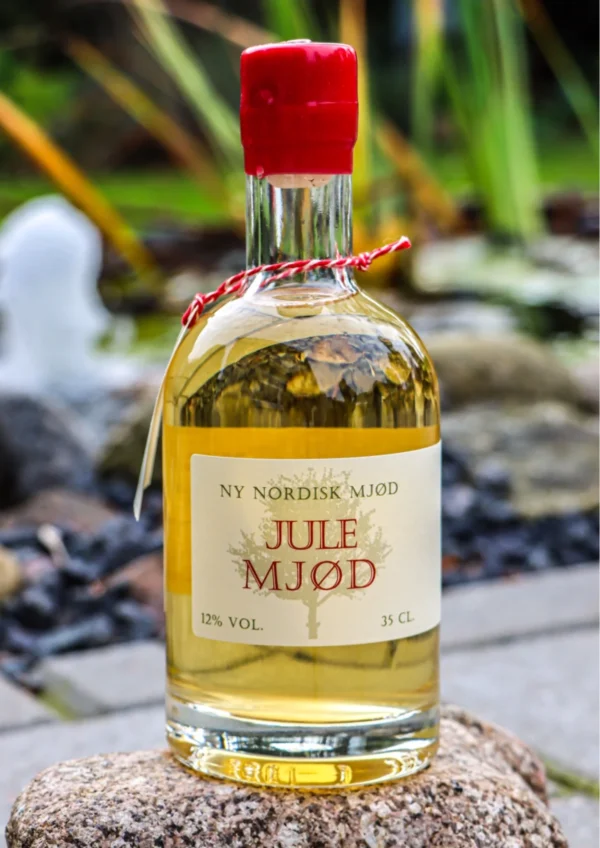 Flaske med julemjød, 35 cl, med rød forsegling placeret på en sten udendørs ved en vandfontæne.