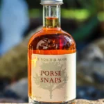 En flaske Porse Snaps på 5 cl med ravfarvet væske stående udendørs på en sten med sløret grøn baggrund