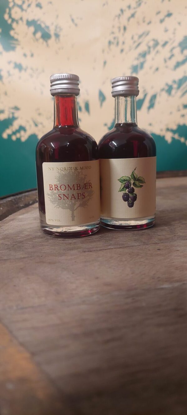 To små flasker 'NY NORDISK MJØD BROMBÆR SNAPS' på 5 cl med mørk væske, stående på en træoverflade.