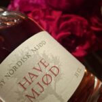 Nærbillede af en flaske 'Ny Nordisk Mjød HAVE MJØD' på 35 cl med en rødlig væske, foran røde blomster.