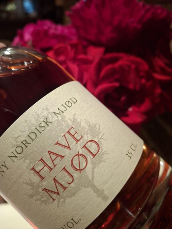 Nærbillede af en flaske 'Ny Nordisk Mjød HAVE MJØD' på 35 cl med en rødlig væske, foran røde blomster.