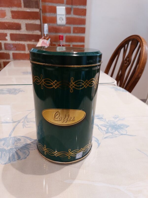 En høj, grøn retro kaffedåse med gulddetaljer og ordet 'Coffee' på en gylden plakette, stående på et blomstret bord.
