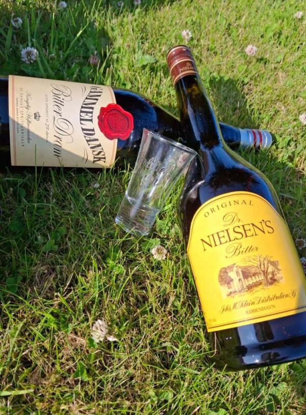 En porcelænsfigur af en beslagsmed og to flasker Gammel Dansk og Dr. Nielsens Bitter, ligger på græsset til en picnic.