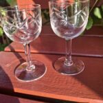 To faset slebne vintage glas på fod, med fine mønstre, placeret på en træbænk i sollys med grønne blade i baggrunden.