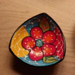 Nærbillede af en farverig, håndmalet tapasskål med et blomstermotiv i rødt, gult og blåt, på et træbord.