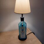 En Bombay Sapphire gin flaske lampe med hvid skærm, står på et lyst træbord med en ledning bagpå.