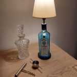 En Bombay Sapphire gin flaske lampe, krystal karaffel, proptrækker og målebæger arrangeret på et lyst træbord.