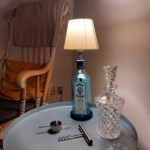 En Bombay Sapphire gin flaske lampe på et lyseblå bord, ved siden af en krystal karaffel, proptrækker og målebæger.