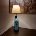 En Bombay Sapphire gin flaske omdannet til en lampe med en hvid lampeskærm, står på et mørkt bord i et hyggeligt rum.