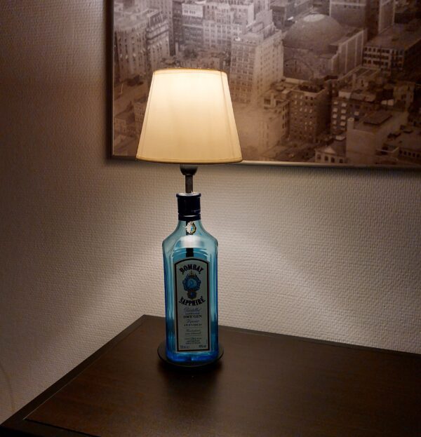 En Bombay Sapphire gin flaske omdannet til en lampe med en hvid lampeskærm, står på et mørkt bord i et hyggeligt rum.
