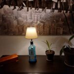 En Bombay Sapphire gin flaske lampe på et mørkt bord, flankeret af to grønne planter. Et sort-hvidt panoramabillede hænger bagved.