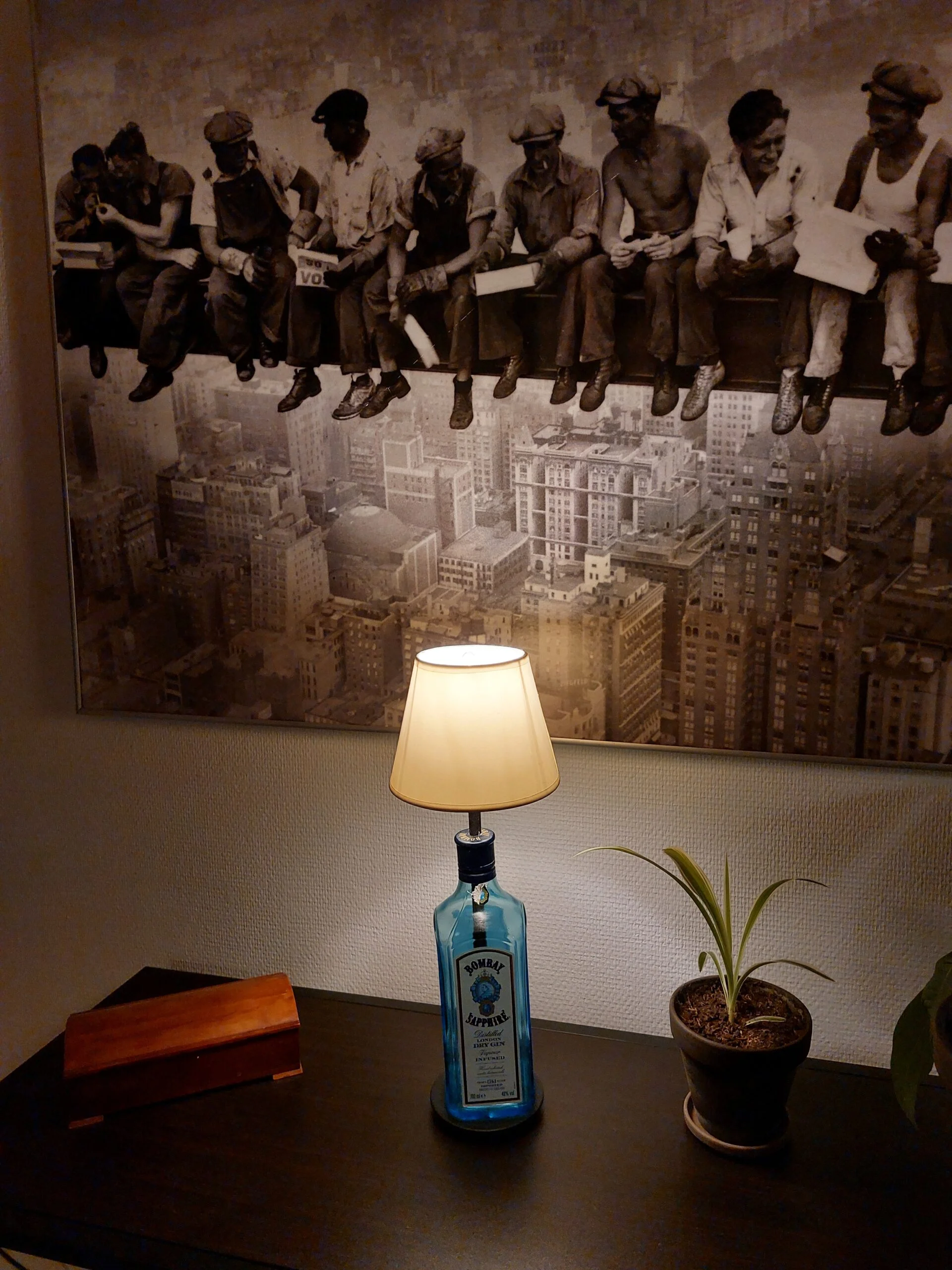 En Bombay Sapphire gin flaske omdannet til lampe med hvid skærm. Står på et mørkt bord med et stort sort-hvidt vægmaleri bagved.