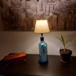En Bombay Sapphire gin flaske lampe med hvid skærm på et mørkt bord, ved siden af en lille plante og en trææske. I baggrunden ses et stort sort-hvidt billede.