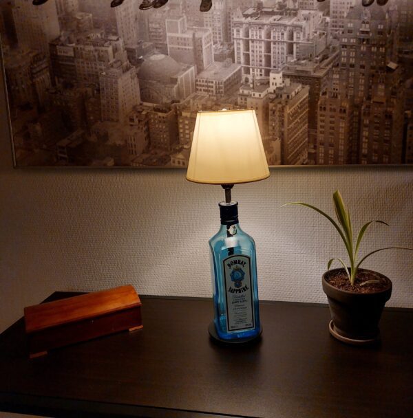 En Bombay Sapphire gin flaske lampe med hvid skærm på et mørkt bord, ved siden af en lille plante og en trææske. I baggrunden ses et stort sort-hvidt billede.