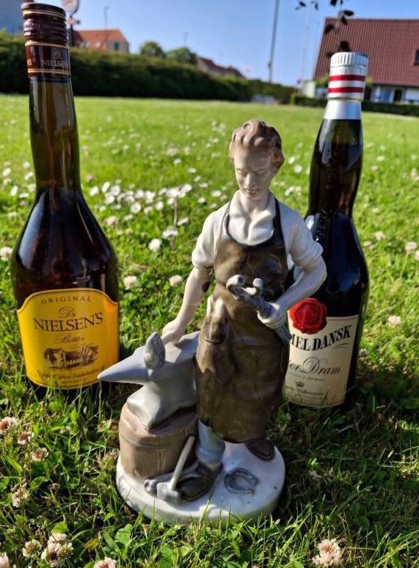 En flaske Dr. Nielsen's Bitter og en keramikfigur af en beslagsmed ved en ambolt, står i grønt græs med hvide blomster.