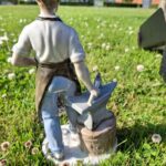 En beslagsmed figur i hvid porcelæn med en ambolt og en hammer, står i grønt græs med små hvide blomster. Set bagfra.