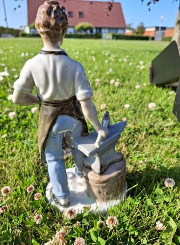 En beslagsmed figur i hvid porcelæn med en ambolt og en hammer, står i grønt græs med små hvide blomster. Set bagfra.
