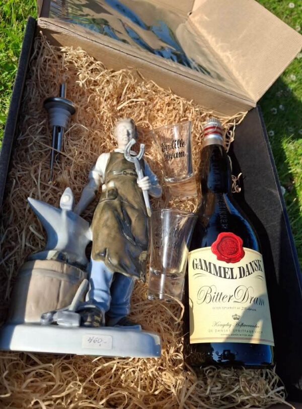 To tomme dramglas med teksten 'En lille dram', placeret i en gavekasse med halm, ved siden af en beslagsmed figur.