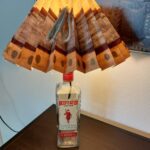 En Beefeater gin flaske omdannet til en lampe, med en plisseret skærm, der lyser varmt. Står på et mørkt bord.