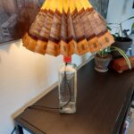 Beefeater gin flaske lampe med strå gul og grå plisseret skærm. Står på et mørkt møbel i et lyst rum.