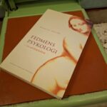 Nærbillede af en bog med titlen 'Fedmens Psykologi' af Annette Ilfeldt, der ligger på foden af en mintgrøn retro vægt.