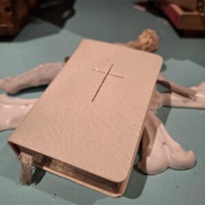 En lille hvid læderindbundet Den Danske Salmebog med et udskåret kors på forsiden, der ligger på en lyseblå overflade.