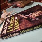 Bogomslag af 'I Herrens navn' af Jack Higgins, der viser en illustration af et kors på en mørk baggrund.
