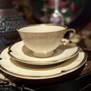 En cremehvid tekop og underkop med elegant guldkant, placeret på en mørk overflade med et brunt, mønstret element i baggrunden.