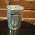 En cylindrisk sparebøsse i metal med stabelbare ringe og en møntindkastsslids, står på sort overflade.
