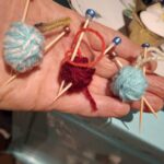 En hånd holder tre håndlavede mini garnnøgler i forskellige farver (blå, rød, grøn) med små strikkepinde i træ og perler.