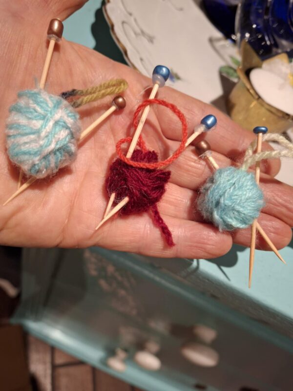 En hånd holder tre håndlavede mini garnnøgler i forskellige farver (blå, rød, grøn) med små strikkepinde i træ og perler.