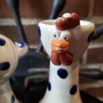 Nærbillede af en hvid keramik hønefigur med blå prikker og orange kam, næb og hagel, stående mod en murstensvæg. Figuren har et åbent hoved som kan bruges til en æggebæger.