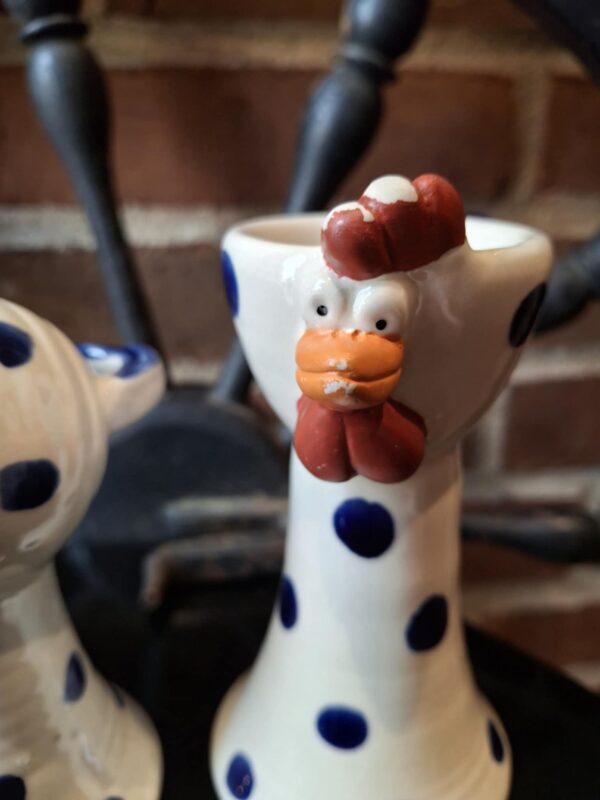 Nærbillede af en hvid keramik hønefigur med blå prikker og orange kam, næb og hagel, stående mod en murstensvæg. Figuren har et åbent hoved som kan bruges til en æggebæger.