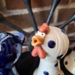 Nærbillede af en hvid keramik hønefigur med blå prikker og orange kam, næb og hagel, stående mod en murstensvæg. Figuren har et åbent hoved som kan bruges til en æggebæger.