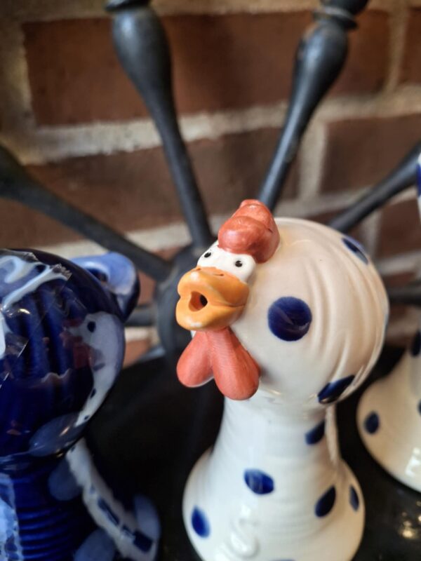 Nærbillede af en hvid keramik hønefigur med blå prikker og orange kam, næb og hagel, stående mod en murstensvæg. Figuren har et åbent hoved som kan bruges til en æggebæger.