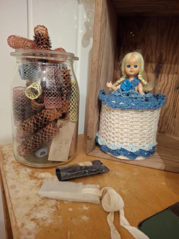 En samling vintage hårfarver og farverige curlere i en stor glasbeholder på et træbord, ved siden af en dukke.