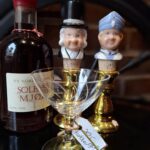 En flaske solbærmjød, et krystalglas, og to vinpropper med håndmalede figurer af H.C. Andersen og Den Standhaftige Tinsoldat står på en mørk overflade.