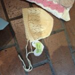 En beige håndstrikket kysehue med hulmønster ligger på et brunt gulv, fastgjort med en sikkerhedsnål.
