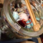 Glasbeholder fyldt med farverige mini garnnøgler og strikkepinde. Et prisskilt sidder på glasset.