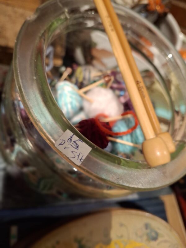 Glasbeholder fyldt med farverige mini garnnøgler og strikkepinde. Et prisskilt sidder på glasset.