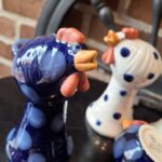To keramik hønefigurer, den ene blå med prikker og den anden hvid med prikker, med et smilende udtryk og orange næb og kam.