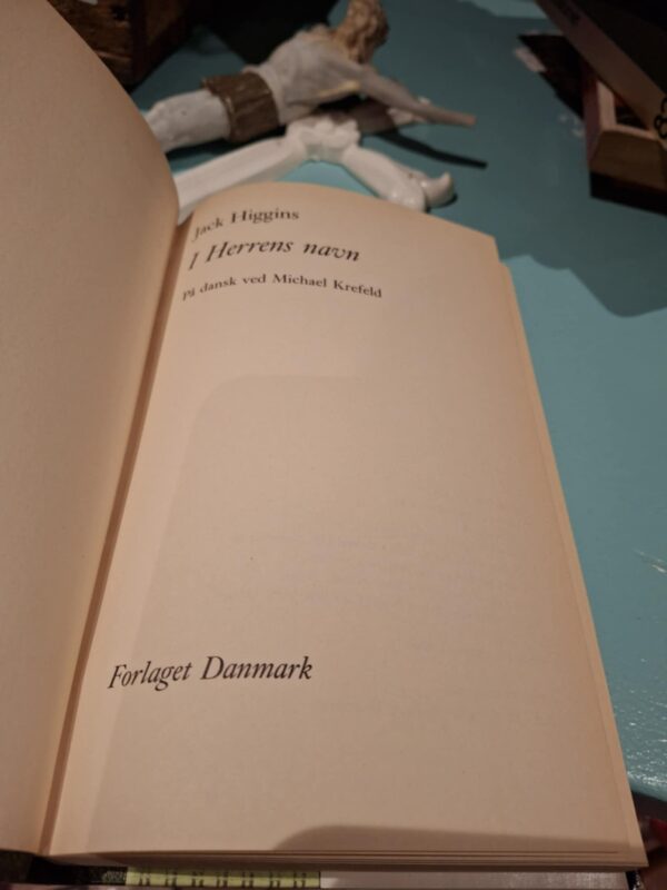Indersiden af bogen 'I Herrens navn' af Jack Higgins, der viser titel, forfatter og forlag 'Forlaget Danmark'.