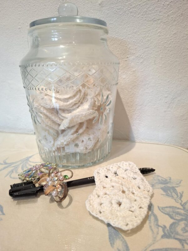 En glasbeholder fyldt med håndhæklede beige make-up fjernerpuder, en makeupbørste og broche på dækket bord.