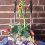 En træblomsterholder med fire glasrør. Tre rør holder tulipaner, to med pyntespyd. En ivy-gren er tilføjet. To fyrfadslys er foran.