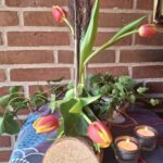 En blomsterholder af træ med fire glasrør, hvori røde og gule tulipaner samt vedbend er arrangeret. To tændte fyrfadslys er i forgrunden.