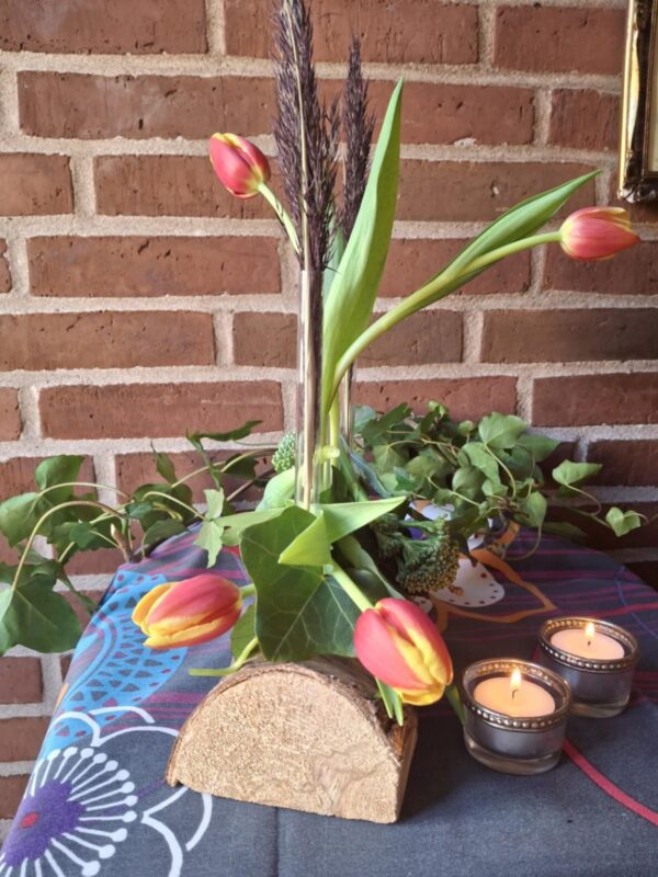 En blomsterholder af træ med fire glasrør, hvori røde og gule tulipaner samt vedbend er arrangeret. To tændte fyrfadslys er i forgrunden.