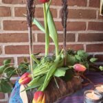 En blomsterholder af træ med glasrør, hvori røde og gule tulipaner samt vedbend er arrangeret. To tændte fyrfadslys er foran. Baggrund murstensvæg.