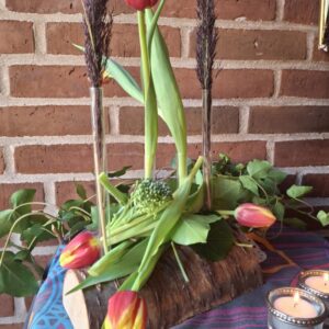 En blomsterholder af træ med glasrør, hvori røde og gule tulipaner samt vedbend er arrangeret. To tændte fyrfadslys er foran. Baggrund murstensvæg.