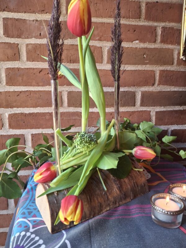 En blomsterholder af træ med glasrør, hvori røde og gule tulipaner samt vedbend er arrangeret. To tændte fyrfadslys er foran. Baggrund murstensvæg.